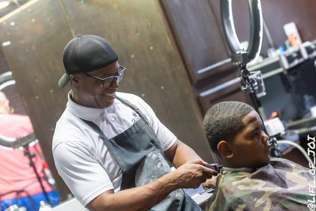 A barber in a flat cap and apron edges a teenager’s lineup at the 888 Fresh Start back to school event.