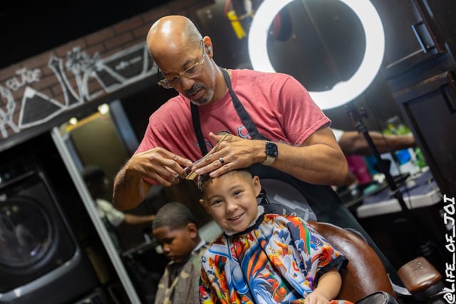 A young boy grins in the barber chair while a barber works on his fresh cut.