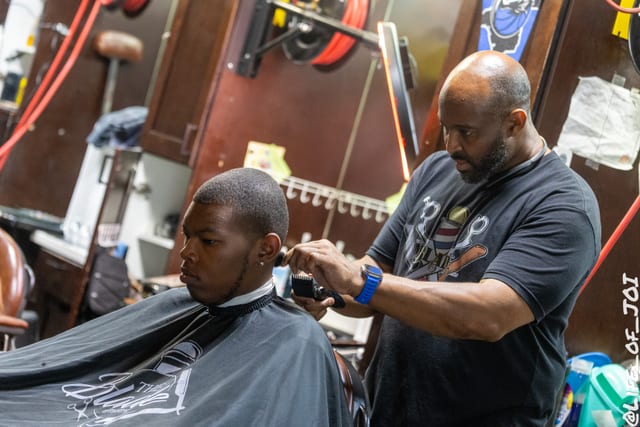 A fade takes shape as a barber works on a student wearing a Blade Barber Lounge cape.