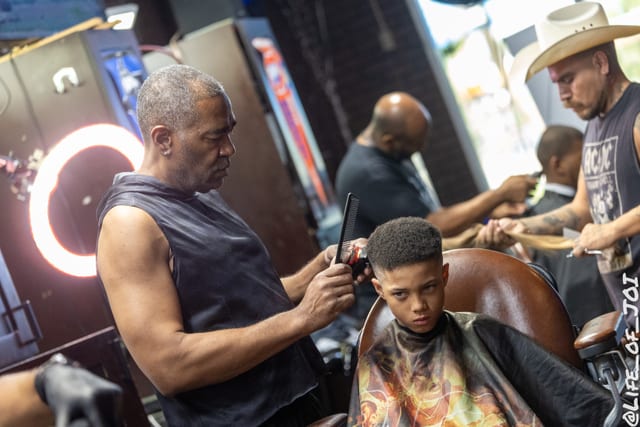 A barber cuts a young boy’s hair with every chair in the shop full behind him.