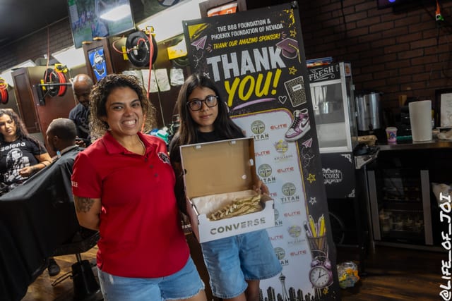 A volunteer in a red polo stands with a student holding her new Converse box at the sponsor banner.