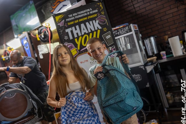 Two students smile with a new teal backpack in front of the 888 Fresh Start Experience banner.
