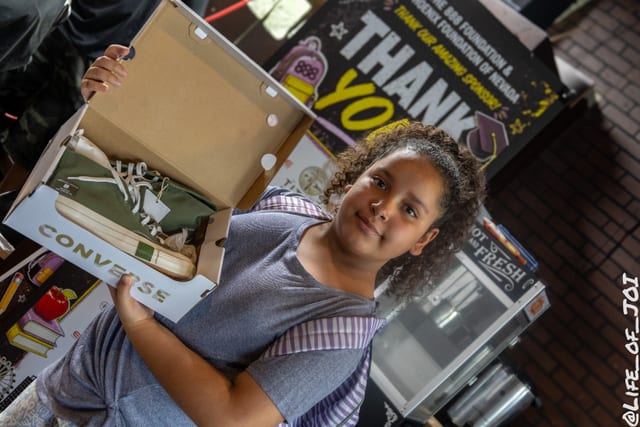 A girl holds up her open box of new sneakers in front of the sponsor banner.