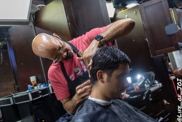 A barber in a work apron shapes a client’s hair in front of the shop mirror.