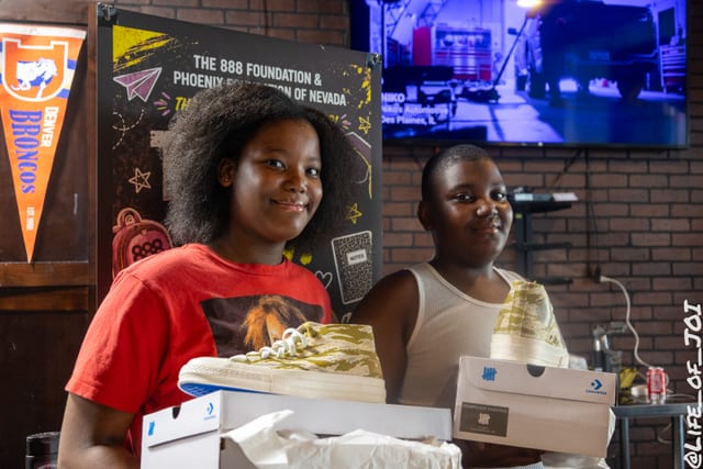Siblings hold up their gold Converse sneakers in front of the 888 Foundation banner.