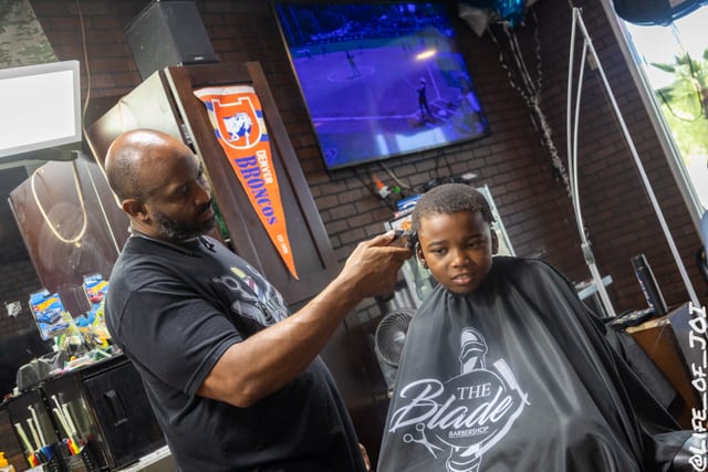 A young boy sits still in a Blade cape while a barber works on his back to school cut.